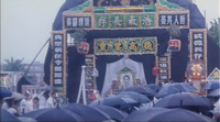 Black umbrellas line up towards very large arches of bright calligraphy and funeral altar.