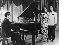 A black and white photograph of Toña la Negra performing with Pedro Vargas and Agustín Lara at the piano in the movie Revancha. She wears an elegant gown with butterlies-­on­squares print.