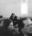 Photograph of a woman standing, speaking, and gesturing pointedly, visible above the heads of seated audience members.