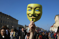 “Save the internet” demonstration in Munich, Germany, in 2019. A Guy Fawkes mask is held aloft by the chin, with demonstrators walking through the street in the background. The mask is yellow. It is a depiction of a smiling man’s face with raised eyebrows, a thin black moustache turned up at the edges, a thin vertical central line of black beard down the chin, red cheeks, and cut-­out holes for eyes.