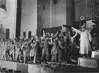 Production photograph depicting a crowd of dockworkers with their arms outstretched and one male actor standing on a wooden crate.