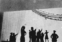 Black-and-white photograph of several actors silhouetted against a lit scrim, as if watching an explosion in the distance.