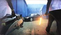 Still from a music video. Two Black men approach a black car with a cutting torch and circular saw.