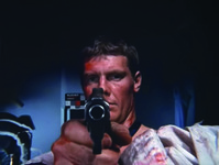 A slightly low-­angle close-­up, with considerably flattened distance, of the bloodied gangster, Chas, looking directly into the camera from center frame. In the extreme foreground, his outstretched arm points his pistol (also) directly into the camera lens. Consequently, the gun barrel appears huge as it takes up the lower center of the frame. In the background, a nondescript grey wall with the shadow of large plant leaves and what looks like a Kleenex dispenser.