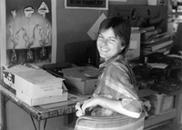 Black-and-white photo. Leighty turns from her work piled on a card table and smiles toward the camera. On the wall are several Chicago Women’s Graphics posters.