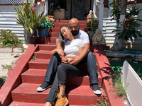 A man sits on red outdoor steps in front of a house on a warm day as he embraces a woman. She looks away. Both wear white T-­shirts and blue jeans.