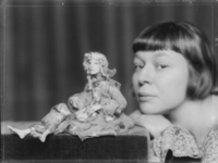 The 1924 black and white photograph of The dollmaker Lotte Pritzel by Atelier Madame d’Ora captures the intimate, almost organic relationship between doll maker and doll by arranging them in a manner that emphasizes the resemblance between both. Set against a dark background, both are positioned at eye level: Pritzel’s chin rests on her hand, which rests in turn on the corner of a velvet platform, where the doll is seated with its legs crossed. While the ornamental motif on the doll’s garment echoes the arabesque pattern of Pritzel’s lace blouse, the doll’s facial features (the oval of the face, the almond-shaped dark eyes) resemble Pritzel’s own.