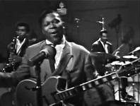 Figure 7. Blues musician B. B. King, dressed in a suit and fronting a full band, sings at a microphone with his eyes closed and without playing his guitar.