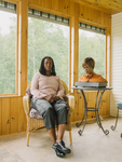 Photo: A Black woman with dark-chestnut skin sits in a wicker chair beside a robotic bust of a Black woman with dark-taupe rubber skin. The robot, which is physically a head and shoulders on a pedestal, sits on a wrought-iron table with a glass top. They are situated in a screened-in sunporch made of light-brown wood. Green foliage is  visible in the background.