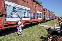 A woman stands in front of the side of a train car holding a photograph of her daughter. A large image of the same woman, holding the same photograph, is affixed to the side of the train car. Many photojournalists are simultaneously taking photos of the woman.