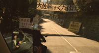 Signs with crude writing line the street as the main character drives to work. One says, "Go to hell, Big-Sato."