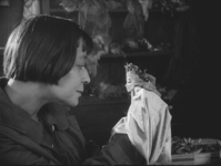 A still shot from the 1923 short documentary film The Pritzel-Doll (Die Pritzelpuppe), commissioned by the UFA film company presents the doll maker Lotte Pritzel at work in her atelier. In the photograph, Pritzel, clad in a work coat, is shown in profile, holding a doll in her left hand and a brush in her right, her gaze directed at the doll’s head, where she is about to paint the mouth. The gaze is intense, intimate, and seems to go both ways: the two heads are shot in perfect symmetry, and the doll seems about to speak to its creator.
