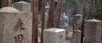 A woman kneels in a graveyard in the background, with calligraphy covered tombstones and markers in the foreground.