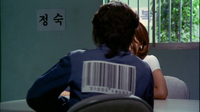 A prisoner meets with a woman in a visiting room with a barcode with numerals printed on the back of his jumpsuit and a paper with black calligraphy posted to the wall.