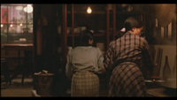 Image of the backs of two people standing in a restaurant, both wearing plaid. In the deep background there are pink and red banners on the wall with calligraphy on them.