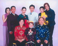 A portrait of the Xiao family. Yunchu, Yuntian, and Yunshu (from left to right) and their partners stand in the back row, with Mr. and Mrs. Xiao seated in front of them with Yunchu’s biracial son standing between them. The older couple and child all wear traditional Chinese dress. Yuntian’s wife, Yanji, wears a Korean-­style dress, while Yuntian and Yunchu’s Malaysian husband are both in business attire. Yunshu and her Australian husband are dressed casually.