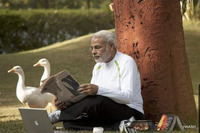 A man, who appears to be Narendra Modi, sits on the ground in a white shirt with his back against a tree and reads a newspaper. A couple of books are opened and turned upside down in the grass next to him alongside an Apple laptop, and two white geese are standing in the background.