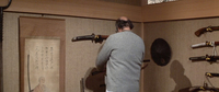 A man examines a sword in a room with many swords and a calligraphy scroll hanging on the walls.