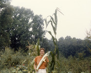 Color photo. Part of the garden surrounds Cohen and a corn stalk towers over her. Forest visible in background. The cucumber is over two feet long.