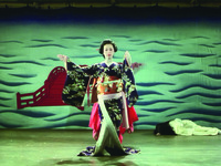 In front of an arresting painted backdrop of a red bridge floating in a vast blue sea, Shinako appears on stage in kimono in the appearance of a giant, stiff bunraku puppet, her arms and whole body manipulated by an unseen puppeteer, concealed in red garb and operating behind Shinako. In the Left corner the seemingly dead body of the child “snow queen”.