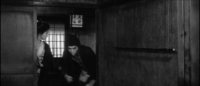 A man enters a room with a lantern with black calligraphy printed on it hanging above him, in black and white cinematography.