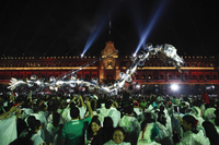 Nighttime revelers, some with cellphone cameras, watch as a giant silver inflatable puppet passes in front of the Palacio Nacional.