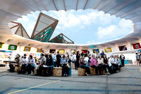 Color image of circular open-­air structure. People of all different ages are sitting on plywood boxes under the open-­air portion of the structure. Some are wearing headphones. They gaze at television screens that ring the interior of the structure.