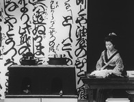 A film still of a woman wearing a kimono seated behind a short table, alongside a wood-burning stove. Behind her, to the left, is a wall covered in black calligraphic text.