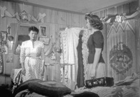 A black and white film still from Konga roja, depicts Marta the Mulata, played by Toña la Negra on the left, and Rosa, played by Maria Antoineta Pons, on the right. In the background are gourds, pictures of saints, and a beaded room divider with the Mexican coat of arms imprinted on it.