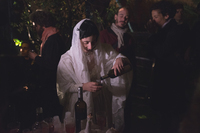 A woman dressed in a wedding dress and veil pours herself a glass of wine.