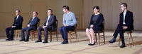A photo of six people wearing formal clothing sitting on evenly spaced wooden chairs in a line in the middle of beige room. In this photo every person is white.