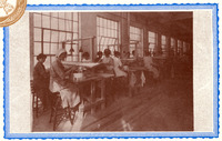 Inside a factory, women add glaze to ceramic spark plugs. Two to four women per work bench sit on tall stools in a long row, some with head covers. Two look toward the viewer.