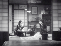 Two women sit on the floor around a short table, with shelves behind them. White calligraphy is painted vertically along the room's entrance frame.