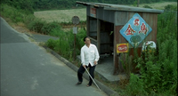 Kikujiro stands in front of a bus stop with red calligraphy printed on it.