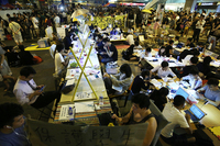 In the study area created on the street, students sit at long tables, some made out of boards placed on top of barriers. Students are using laptops, tablets, phones, and printed texts. In this night-time photo, lights glow over the desks. On either side of the street, crowds pass by, with some stopping to look at the students