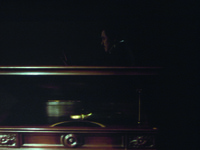 In the background, with only a sliver of light from off-­screen Left, Aochi in profile moves cautiously through the dark, with his hand in front of him. Foreground, in extreme close-­up, a seemingly massive phonograph player, with decorative wood paneling and shiny brass needle. It is better lit from off-­screen Left and front.