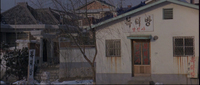 Outside image of a house in winter. The house has rust stains and two signs that feature black and red calligraphy. The door also features red calligraphy on its windows. To the left of the house, a white sign posted in the ground has black calligraphy.