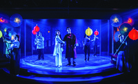 A color photo of a scene on stage. Two figures locking arms are standing at the center, both gazing up to the sky. Seven figures are standing around them, with six of them each holding a lantern on a long stick with both hands and the seventh figure holding a baby in her arm at the left corner of the stage.
