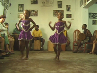 An still image from film showing young girls performing traditional Afro-­Peruvian dance at Ballumbrosio family studio in a room surrounded with photos from family history.