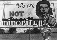 A black and white photograph of a city mural with a male Chicanx activist pointing at “you”; in front of it, a child behind a chainlink fence.