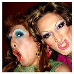 Two persons look at the camera, gesturing with open mouths. They wear lipstick, glittery eyeshadow, big eyelashes, wigs, and long earrings.