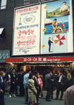 A large billboard for a Korean Film Festival with many people on the busy street looking up towards it.