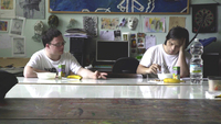 Color screenshot from a documentary film showing two adults with Down syndrome seated and eating at a narrow table, 1facing the same direction about one meter apart from each other.