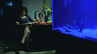 A shot from Visage showing a woman reading at a table on which sits a picture of a deceased woman. Next to the table is a big fish tank.