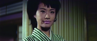 Close-­up of Hanako, center, from Fuyu’s point of view and in front of the striped partition wall. She sizes him up with a hard-­nosed, contemptuous expression. The candy-­striped pattern on the partition resembles that on her kimono, though a lighter shade. Where it ends, screen Left, we see some of the depth of the purple-­wallpapered room behind it.