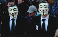 Anti-­ACTA demonstration in Frankfurt am Main, Germany. Two men are pictured at a demonstration, both wearing white Guy Fawkes masks. The mask depicts a smiling man’s face with raised eyebrows, a thin black moustache turned up at the edges, a thin vertical central line of black beard down the chin, red cheeks, and cut-­out holes for eyes. Both men are wearing standard business attire: black suits with white shirts and black ties. The man on the right has a sticker of the Guy Fawkes mask in black and white on his chest, below the lapel.