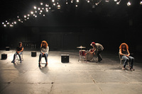 Three actors sit in folding chairs on a darkened stage, their faces barely visible. Behind them someone sets up a drum kit.