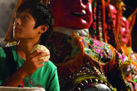A shot from God Man Dog showing a young boy eating a bun in front of a huge puppet of Nezha, a Taiwanese religious figure.
