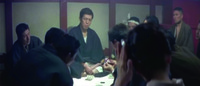 In the midst of the card game, Okaru-­Hachi is seen in the background (in front of the shōji), Left of center and below eye-­level, looking into the camera. Right of him, extreme foreground is the back of a woman’s slightly inclined head and neck, with her hand (seemingly enlarged by the wide-­angle) appearing to smooth her hair. The gamblers make up the middle ground between them filling the area Left and Right of Okaru-­Hachi.