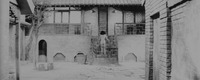 Long Shot of a traditional Chinese brick courtyard stretching across the background, the walls of its entranceway framing the Left and Right foreground. There are patterned doors, odds and ends, a lone tree. Harumi is seen from behind, racing up ten stone steps (center Right) leading to the landing of a house. She easily sheds her kimono, stark naked as she rushes through the Left of two doors. The frame is artificially brightened by an excess of white light.